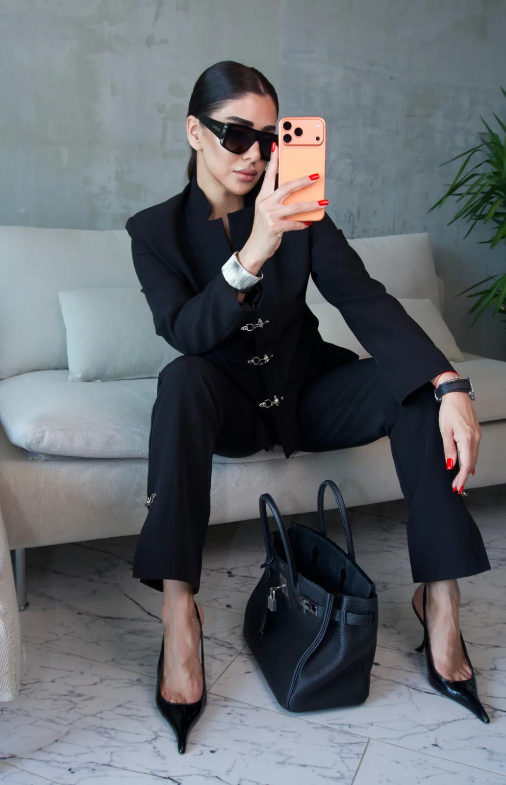 Black stand-collar suit, model seated on a sofa taking a photograph on a phone